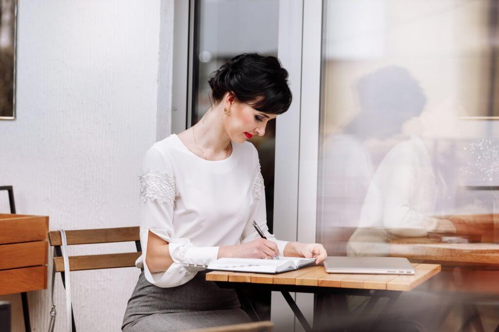 Serious focused smart businesswoman is writing notes on notepad, making agenda on personal organizer
